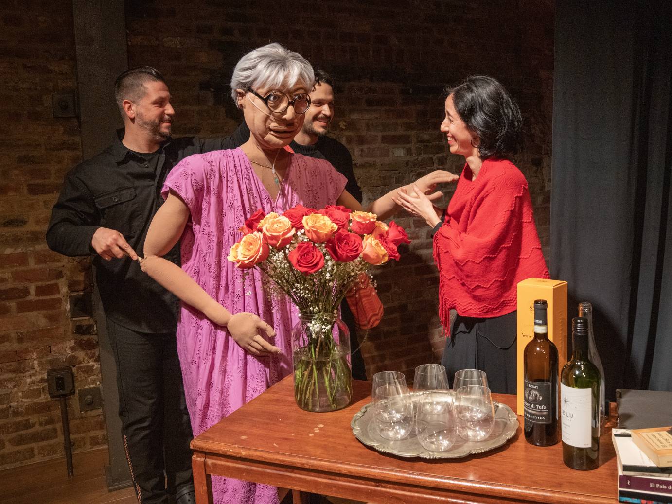 Sonia, the puppet, dressed in a pink frock, gray-haired wears dark framed round glasses. Her two male puppet handlers and a dark haired woman wearing a red shawl smile at one another. Bottles of alcohol and glasses resting on a silver tray, sit atop a table. a vase of red and apricot roses gather in a vase. Sonia stands in front of the roses